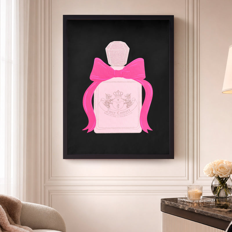 The Shadow Box Wall Art, 18″ x 24″, featuring a fashion-inspired pink perfume bottle embellished with a large pink bow, is displayed elegantly on a beige wall above a table adorned with cream roses and a lamp.