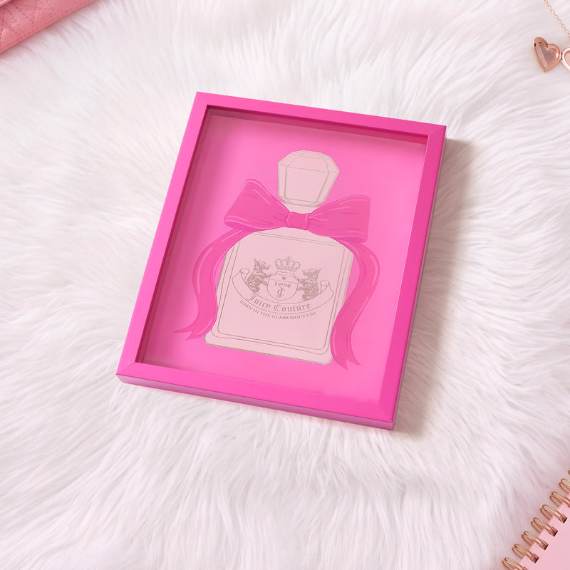 A Juicy Couture Shadow Box Wall Art (12″ x 16″) with a pink frame features a perfume bottle decorated with a large bow, sitting atop a white faux fur surface. Showcasing various shades of pink, this chic, fashion-inspired artwork is complemented by the partial view of a spiral notebook and a heart-shaped paperclip nearby.