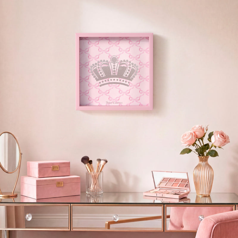 A vanity table with makeup brushes, pink boxes, a mirror, and a vase of pink roses is complemented by the Shadow Box Wall Art (14″ x 14″) hanging above. Featuring a silver crown on a patterned background in a pink frame, this Juicy Couture-inspired piece adds a fashionable accent to the chic setting.