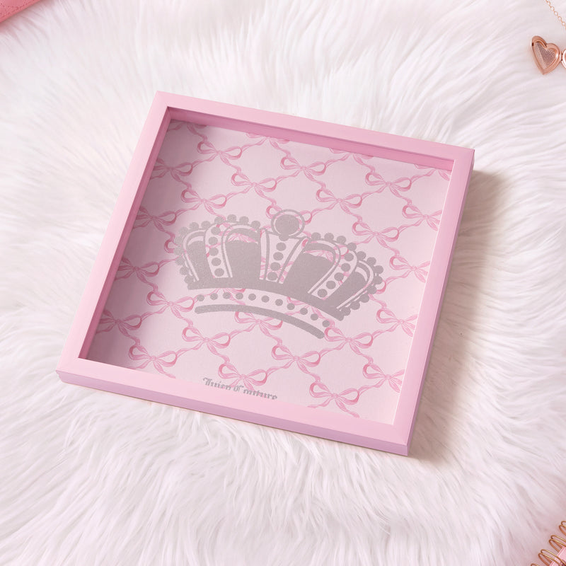A 14″ x 14″ Shadow Box Wall Art with a pink frame, showcasing a pattern of pink bows and a large silver crown design inside, displayed on a fluffy white surface—the perfect Juicy Couture-inspired accent for any space.