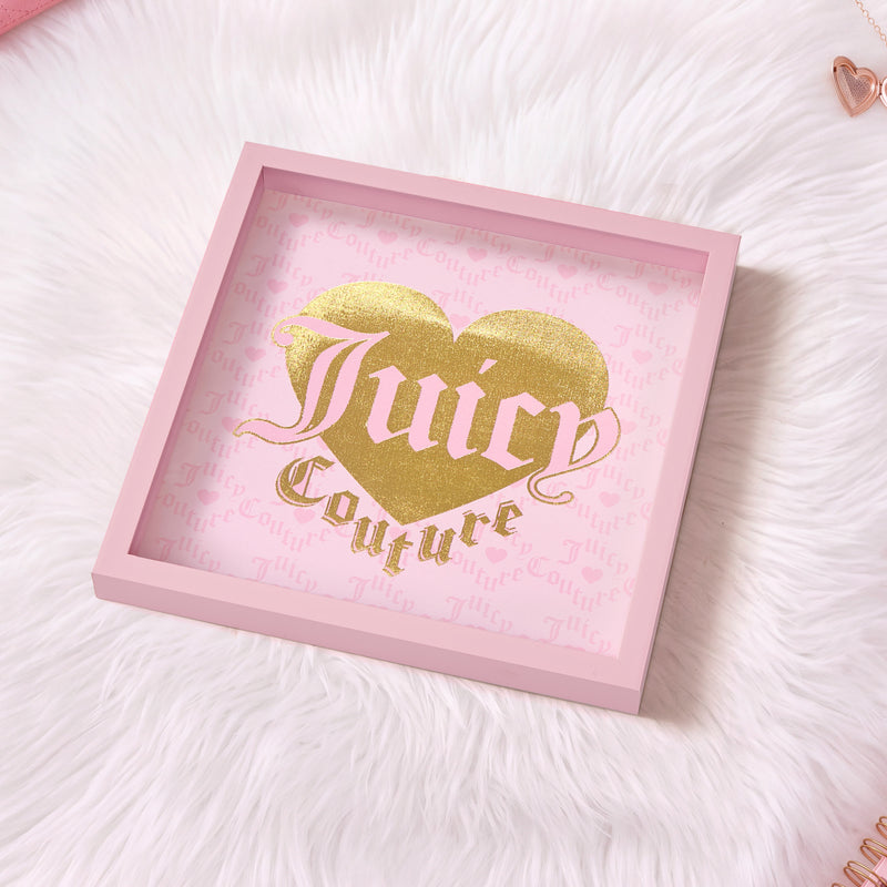 A pink-framed Shadow Box Wall Art (14″ x 14″) featuring a gold heart and “Juicy Couture” in elegant gold ornate letters sits atop a white furry surface, creating a stylish, fashion-inspired statement piece. The background is adorned with light pink Juicy Couture text, making this stunning art the perfect addition to your Juicy Couture wall decor collection.
