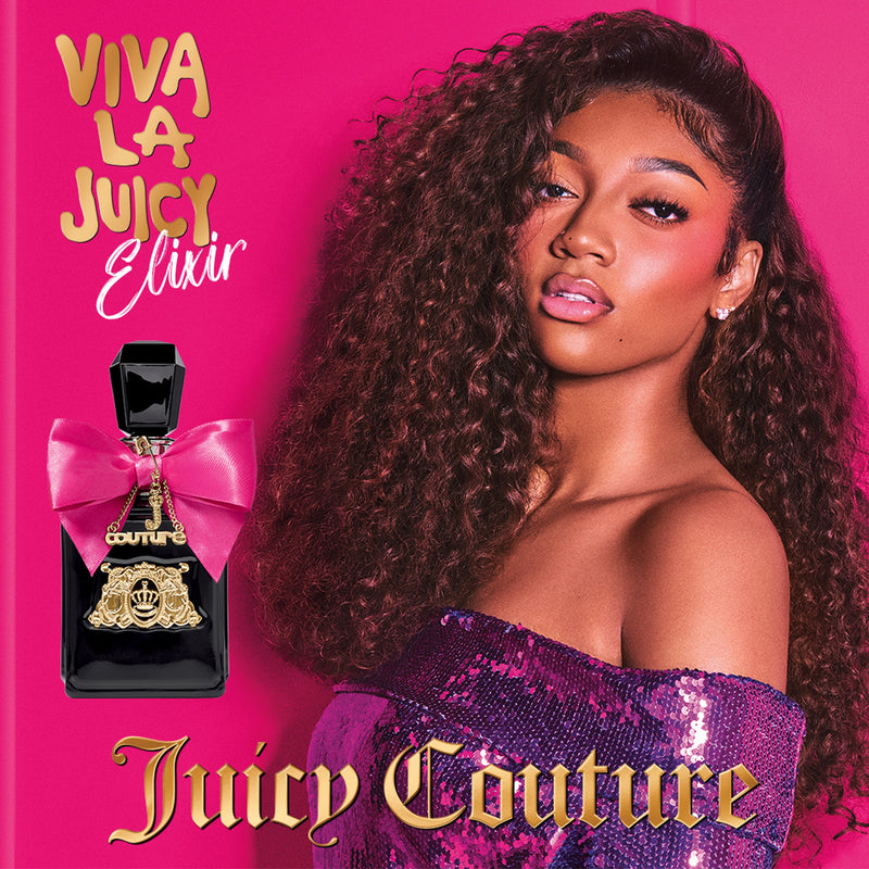 A woman with long, curly hair stands before a pink backdrop in a purple sequin top next to a black bottle with a pink bow. Gold text reads: Viva La Juicy Elixir Eau de Parfum Spray, 3.4 oz—Juicy Couture’s floral amber gourmand.