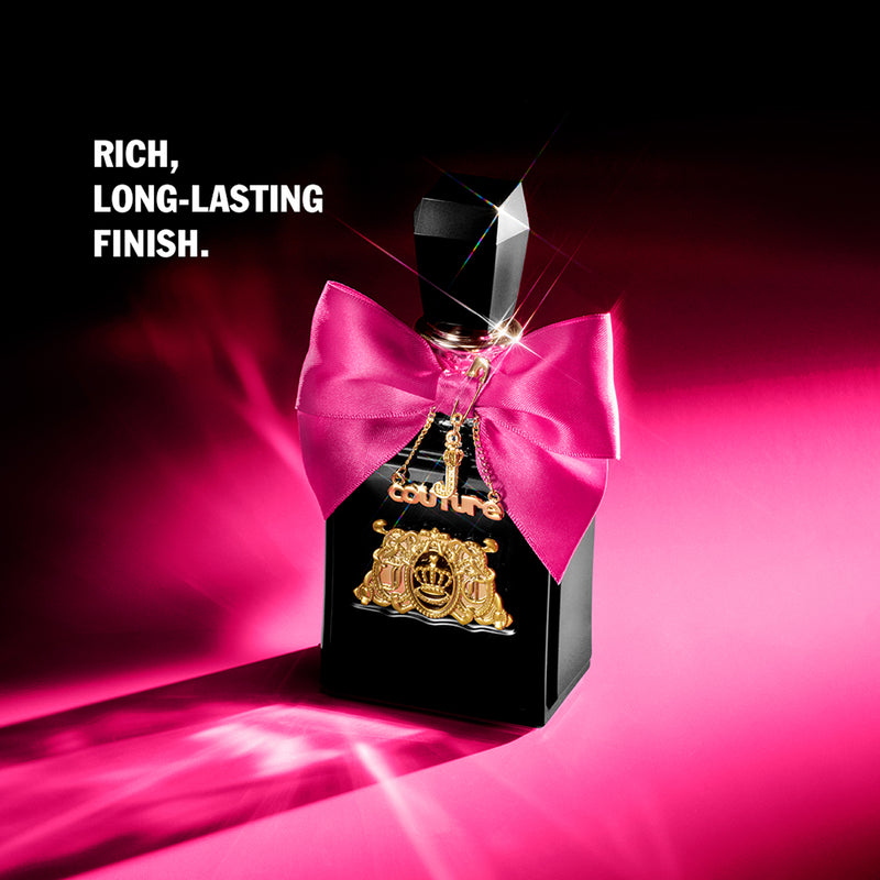 A black Viva La Juicy Elixir Eau de Parfum Spray (3.4 fl oz) bottle, adorned with a pink bow and gold charms, sits on a pink surface under bright light. White text reads: RICH, LONG-LASTING FINISH. Discover its allure.