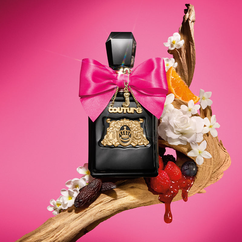 A black Viva La Juicy Elixir Eau de Parfum Spray, 3.4 fl oz bottle with gold accents and a pink bow sits on a twisted wooden branch, surrounded by white flowers, an orange slice, berries, and cocoa beans against a bright pink background.