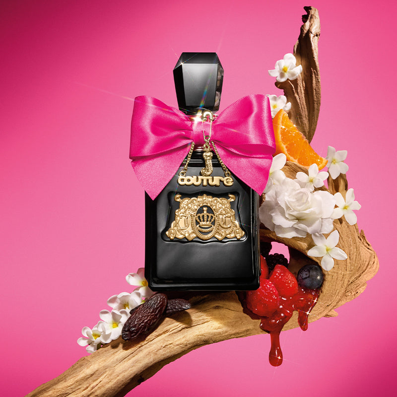 A Viva La Juicy Elixir Eau de Parfum Spray Pen, 0.33 oz, with black and gold accents and a pink bow rests on a wood branch amid white flowers, orange slices, and berries—a vibrant women’s fragrance by Juicy Couture.
