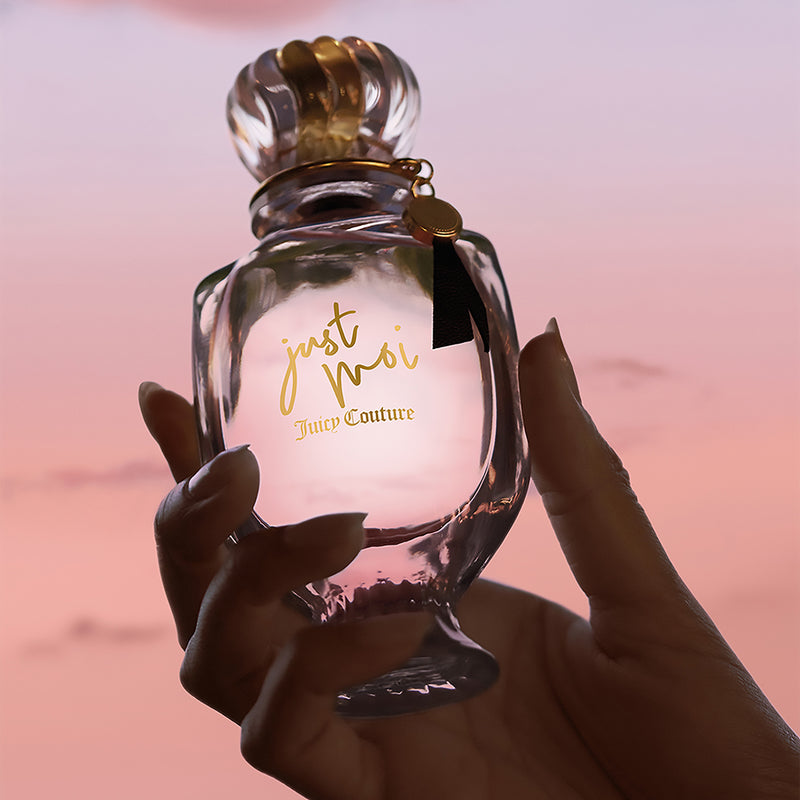 A hand holds a Just Moi 3 Piece Fragrance Gift Set by Juicy Couture against a soft pink and purple sunset sky.