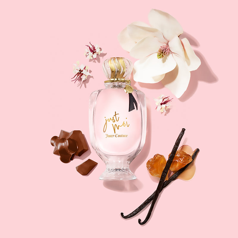 A Just Moi 3 Piece Fragrance Gift Set by Juicy Couture is artfully displayed with chocolate pieces, amber stones, vanilla beans, and white flowers on a pink background, evoking the indulgent and enchanting essence of this feminine Eau de Parfum collection.