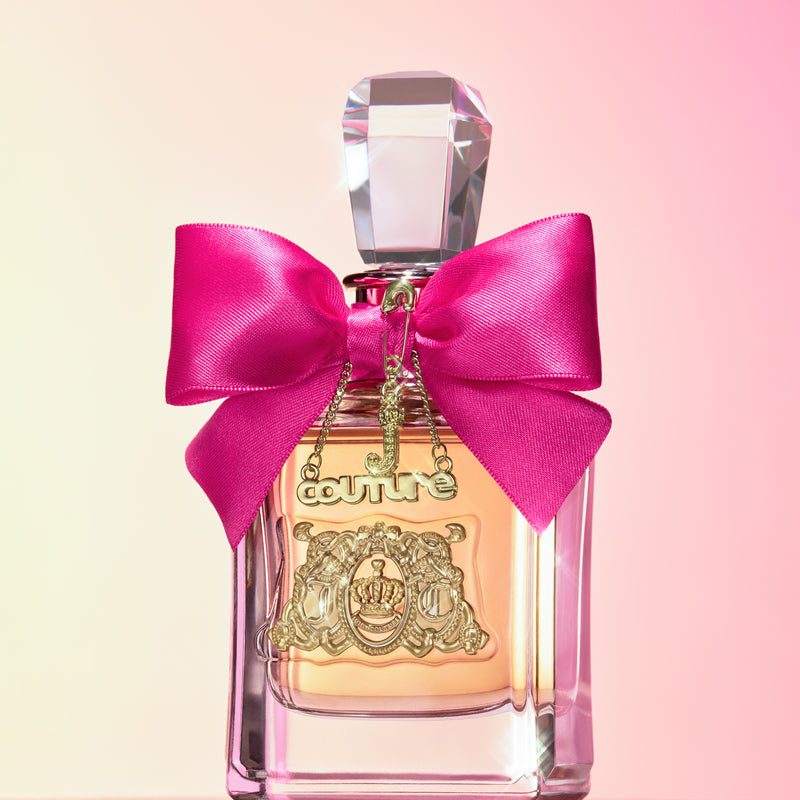 A perfume bottle from the Viva La Juicy 3 Piece Fragrance Gift Set, adorned with a pink bow, gold charm, and clear cap, is displayed against a pink gradient background. The label is embellished with a decorative gold crest, reflecting the playful elegance characteristic of Juicy Couture perfumes—making it an ideal addition to any fragrance collection and capturing the signature essence of Viva La Juicy.