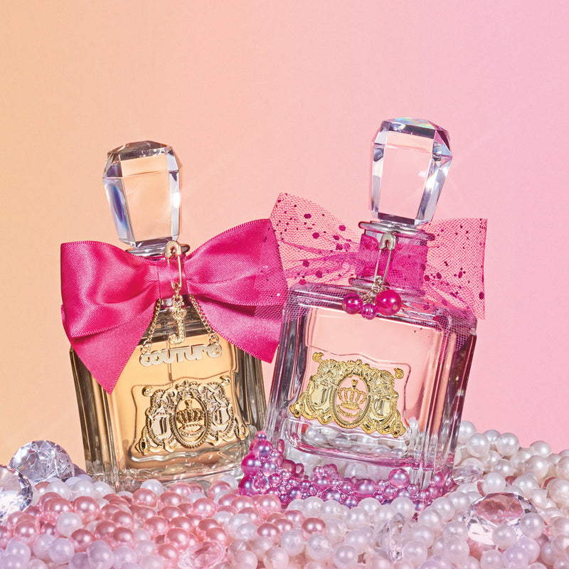 Two elegant bottles from the Viva La Juicy Le Bubbly 3 Piece Fragrance Gift Set, adorned with pink bows and gold labels, are surrounded by pink and white pearls on a pastel gradient background, capturing the playful essence of this signature scent.