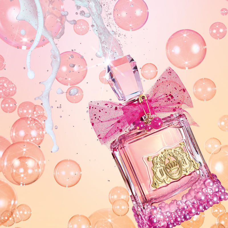 A pink perfume bottle adorned with a jeweled cap, gold emblem, and a pink bow—emblematic of Juicy Couture fragrance—rests amidst floating pink bubbles and white splashes on a gradient peach-pink background, perfectly evoking the playful spirit of the Viva La Juicy Le Bubbly 3 Piece Fragrance Gift Set.