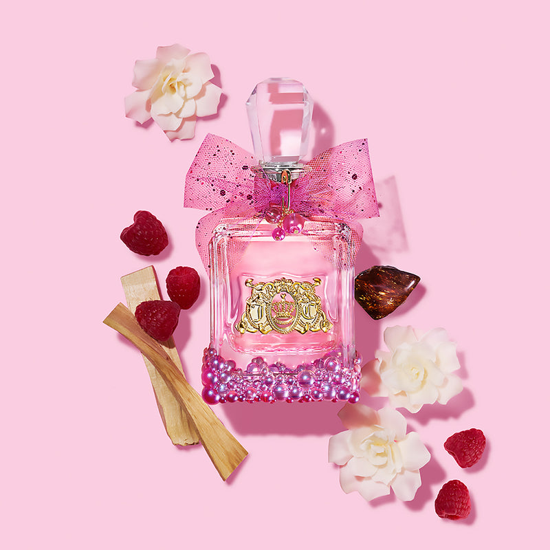The Viva La Juicy Le Bubbly 3 Piece Fragrance Gift Set, featuring a pink perfume bottle adorned with a bow, is beautifully arranged among white flowers, raspberries, pink pearls, wood sticks, and a brown seed on a pastel pink background.
