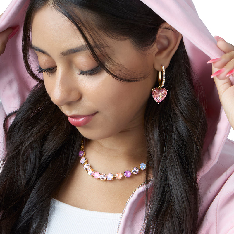 A woman in a pink hoodie looks down, touching her hood. She wears sparkling pink and white gem necklace and Heart Charm Hoop Earrings. Her long dark hair is styled loose.