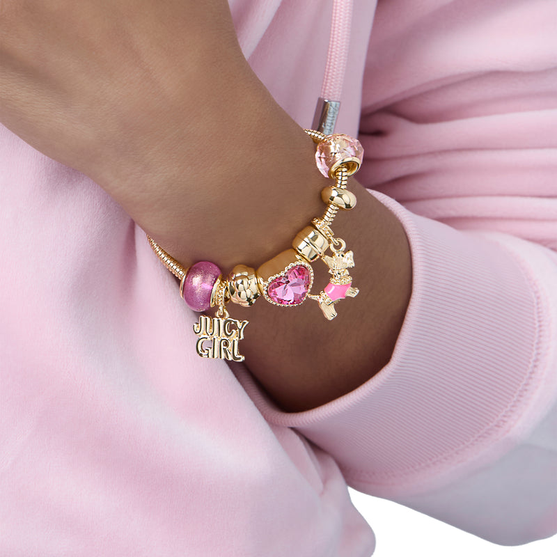 Wearing a pink hoodie, a person displays the Juicy Girl and Scottie Dog Charm Bracelet by Juicy Couture, adorned with pink beads and playful charms like a heart, a bear, the iconic Juicy Girl text, and the signature Scottie Dog—an ideal statement piece for any Y2K jewelry enthusiast.