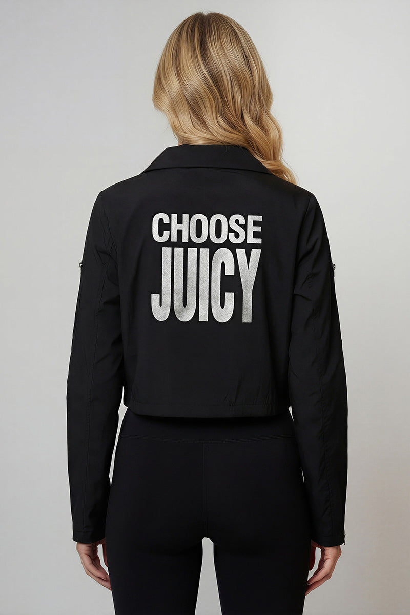 A woman with long blonde hair is seen from the back, wearing the Asymmetrical Moto Windbreaker in black crinkle nylon with large white CHOOSE JUICY text, paired with matching black pants.