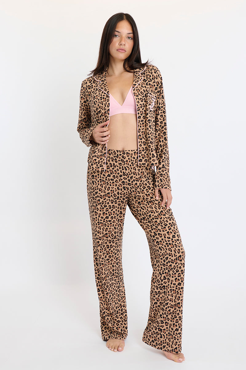 Woman barefoot against a plain background, wearing the Plush 3-Piece PJ Set: a pink triangle bralette with matching leopard print elastic waist pants and a button-down shirt.