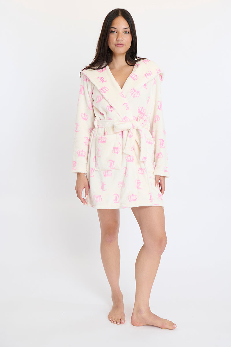 A woman stands barefoot, facing forward in a cream Hooded Luxe Plush Robe with a pink crown pattern. The knee-length, long-sleeved robe is tied at the waist. She poses against a plain white background.