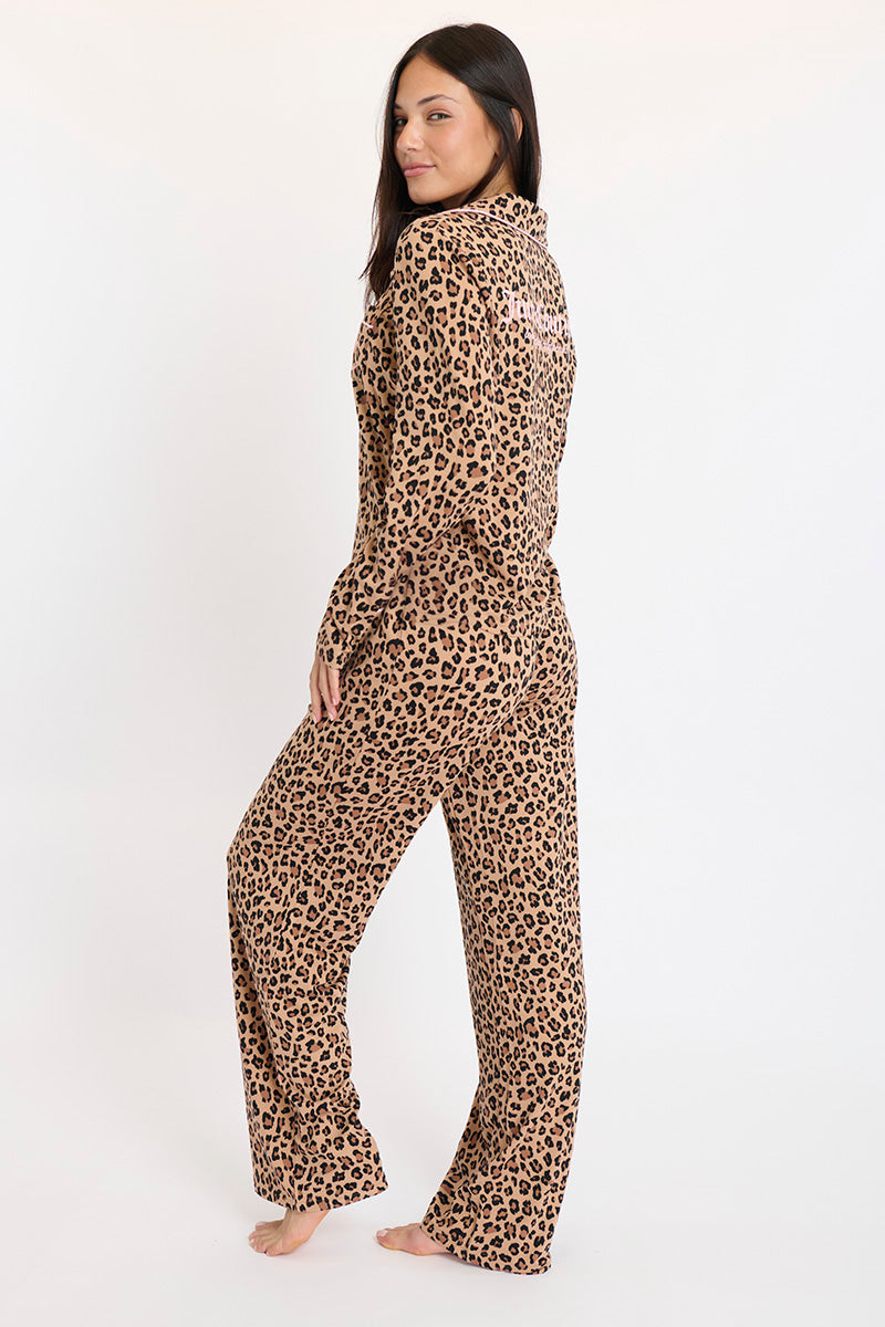 A woman stands barefoot on a white background, modeling the Plush 3-Piece PJ Set in a brown and black leopard print. The button-down shirt and elastic waist pants set is shown as she turns slightly to the side, looking over her shoulder.