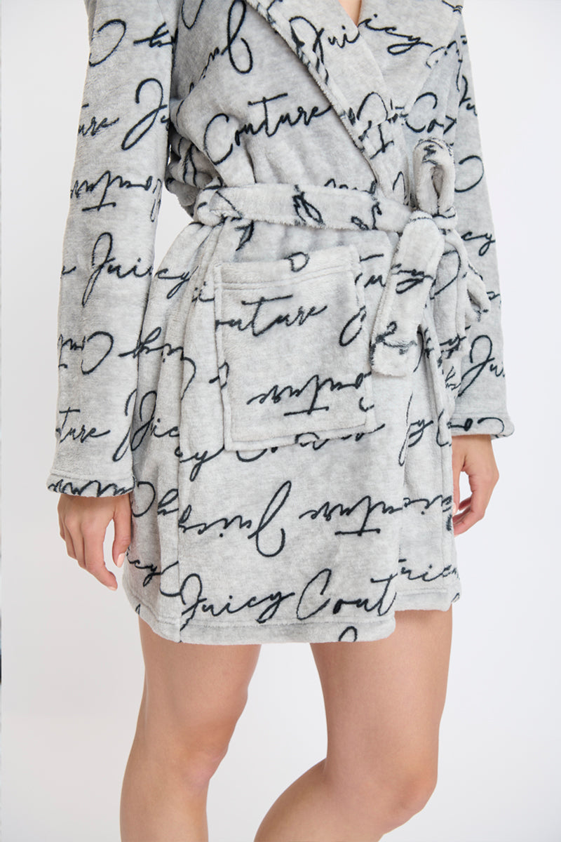 Someone wears a soft, light gray Hooded Luxe Plush Robe featuring black Juicy Couture cursive, long sleeves, a belted waist, and a large front pocket—ideal for cozy self-care moments.