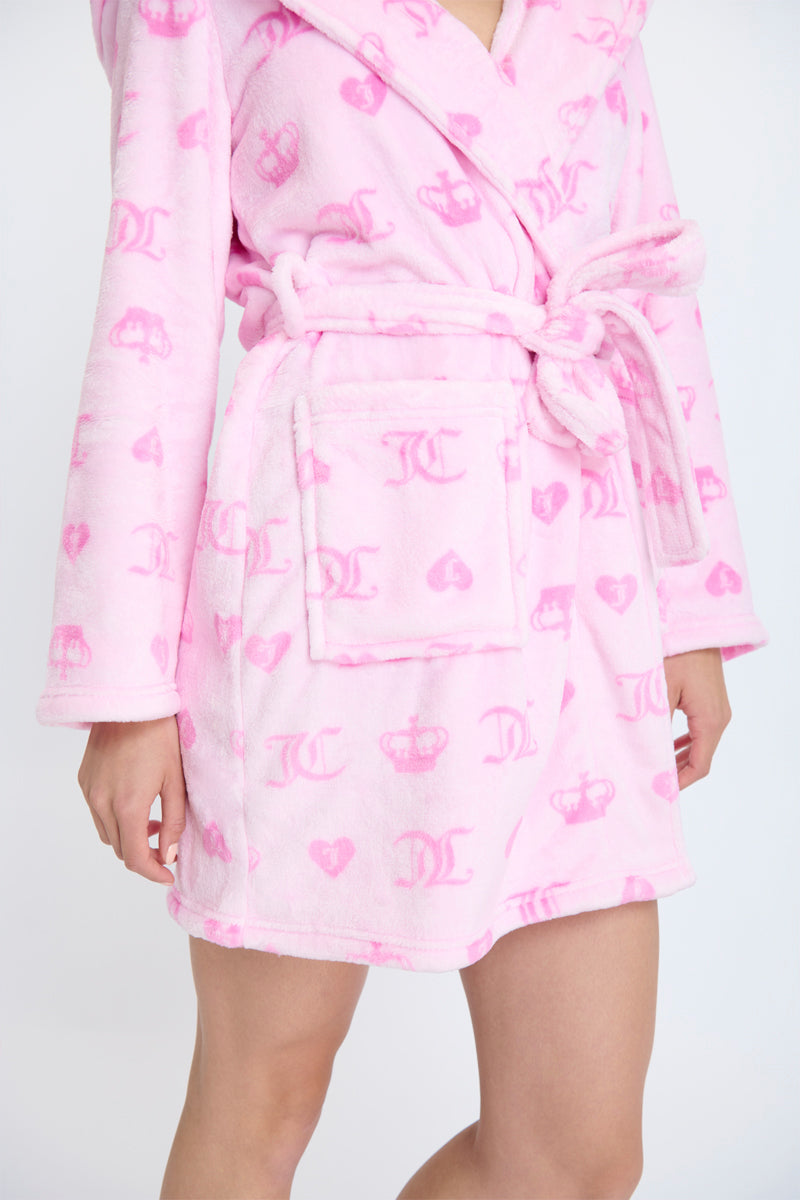 Wearing a Hooded Luxe Plush Robe in light pink with a belt, hood, and an all-over pattern of darker pink crowns, letters, and hearts. This cozy robe is perfect for self-care moments. Model’s face is not visible.
