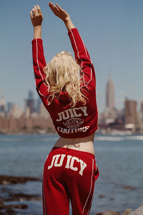 A blonde woman stands with her back to the camera, arms raised toward the sky, wearing a red Scottie Fleece Quarter Zip Sweatshirt with white stripes and a Scottie crest graphic, set against a blurred New York City skyline and river.