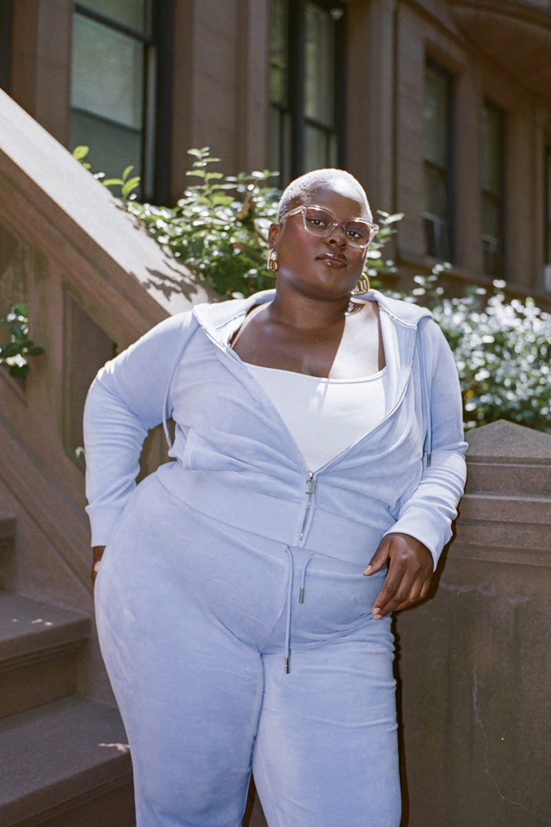 A person in glasses poses outside on stone steps, wearing the Plus-Size OG Big Bling Velour Track Pants in light blue, showcasing a subtle Y2K flair, with greenery and a building in the background.
