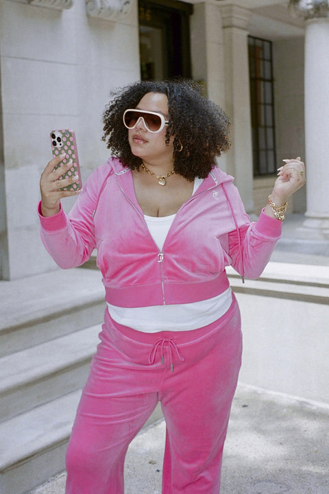 A woman in the Plus-Size OG Big Bling Velour Hoodie, featuring a bold Juicy logo, and large white sunglasses holds a phone while standing confidently on stone steps outside a building.