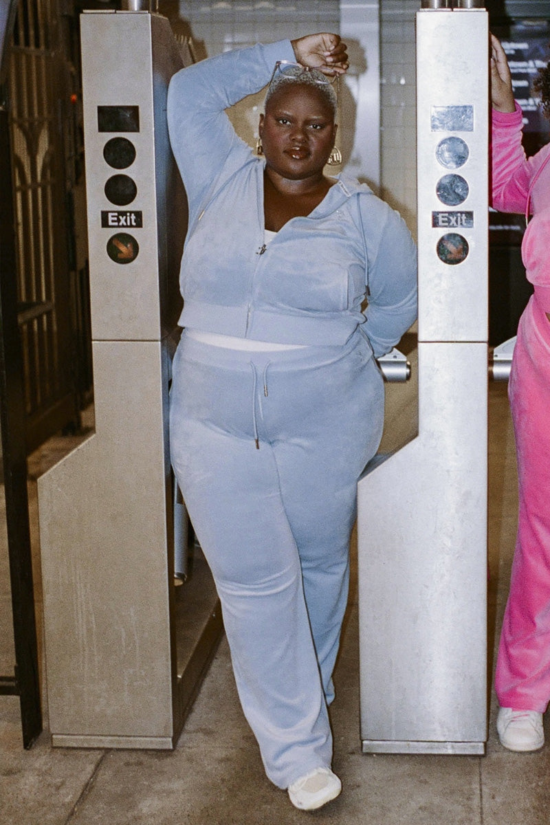 Wearing the Plus-Size OG Big Bling Velour Track Pants in light blue—a nod to Y2K fashion—a person poses confidently between subway exit turnstiles, one arm raised and resting on the metal bar.