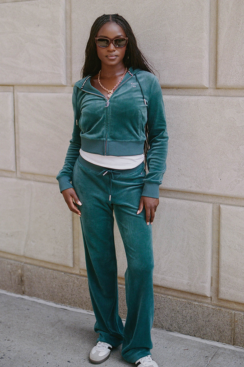 A person stands outside against a stone wall, wearing the OG Big Bling Velour Hoodie in teal with a rhinestone Juicy logo, sunglasses, white sneakers, and layered necklaces.