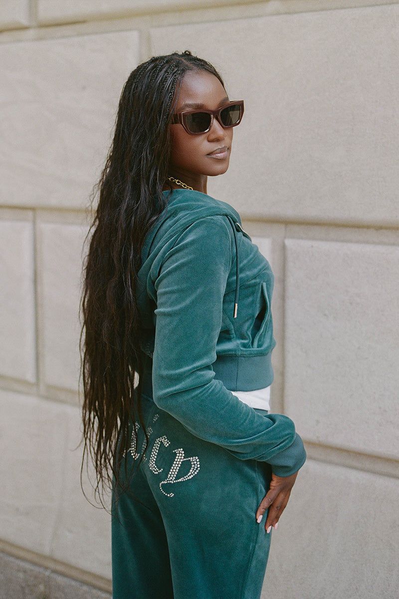 A woman wearing sunglasses and a teal OG Big Bling Velour Hoodie stands in front of a stone wall, looking over her shoulder. The hoodie features a rhinestone Juicy logo on the back, adding a signature Y2K flair. She has long, wavy hair and wears a gold chain necklace.