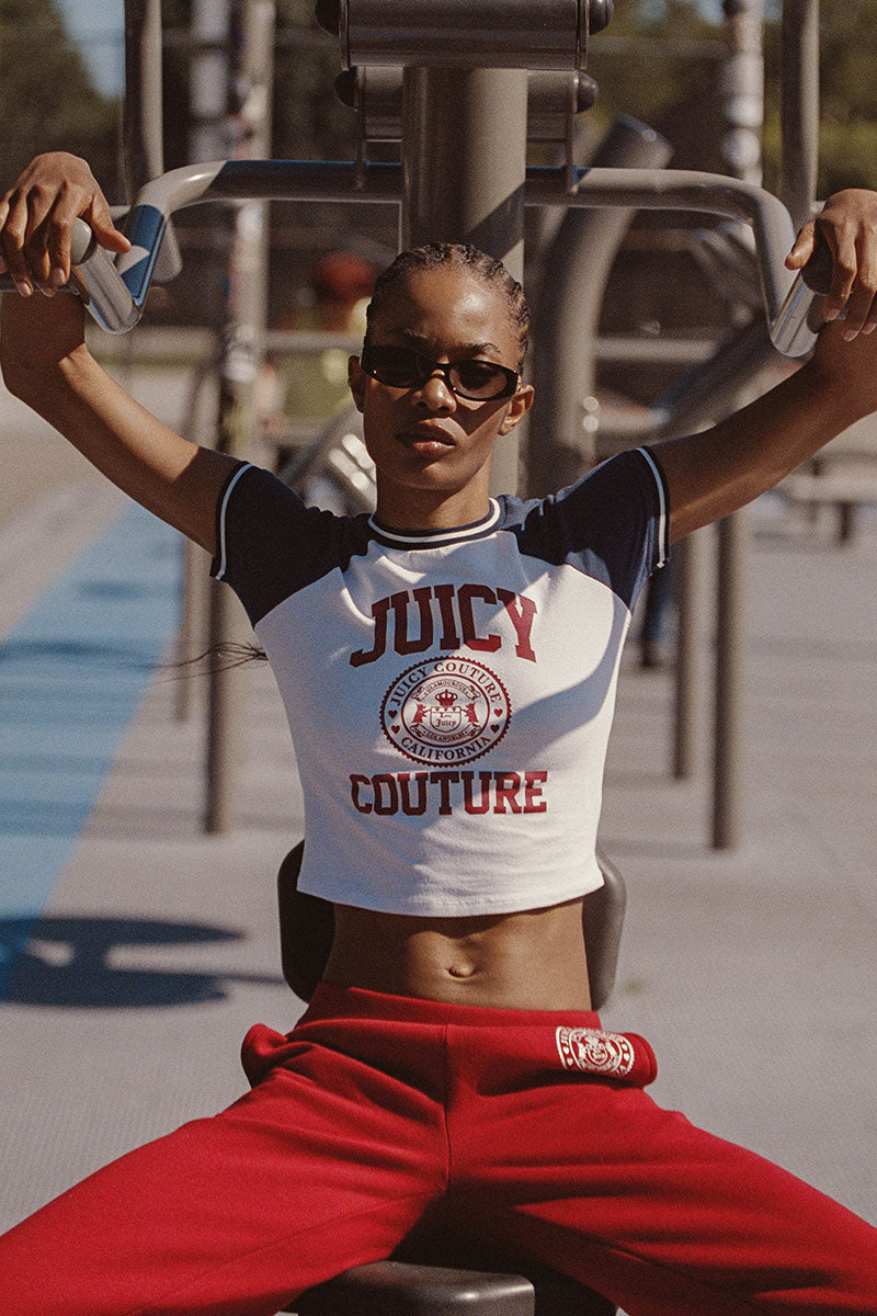 A person wearing sunglasses, the Scottie Baby Tee with bold collegiate graphics, and red sweatpants sits at an outdoor gym machine, gripping the handles and looking confidently at the camera.