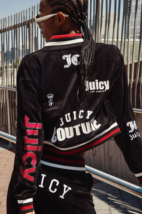 A person with braided hair and sunglasses wears the Juicy Couture Mixed Logo Cotton Velour Bomber Jacket in black, featuring a mix of Juicy graphics for a sporty throwback vibe, while standing outdoors near metal railings.