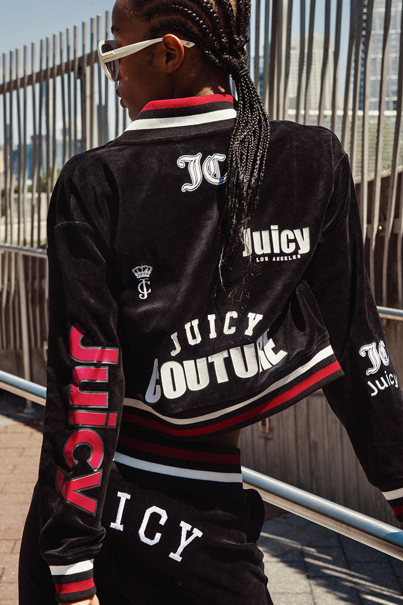 A person with braided hair and sunglasses wears the Juicy Couture Mixed Logo Cotton Velour Bomber Jacket in black, featuring a mix of Juicy graphics for a sporty throwback vibe, while standing outdoors near metal railings.