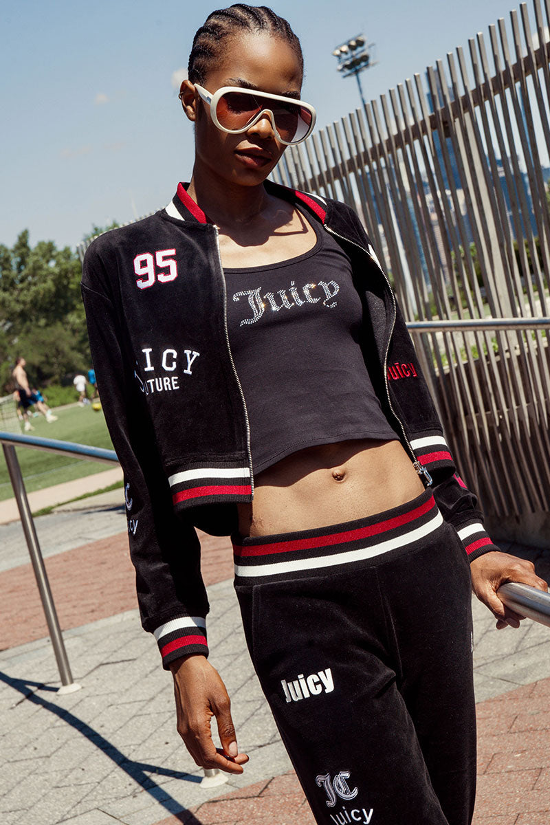 Wearing the Mixed Logo Cotton Velour Bomber Jacket in black and sunglasses, a person poses outdoors near a metal railing, channeling sporty retro vibes with bold Juicy graphics as trees and a sports field appear in the background.