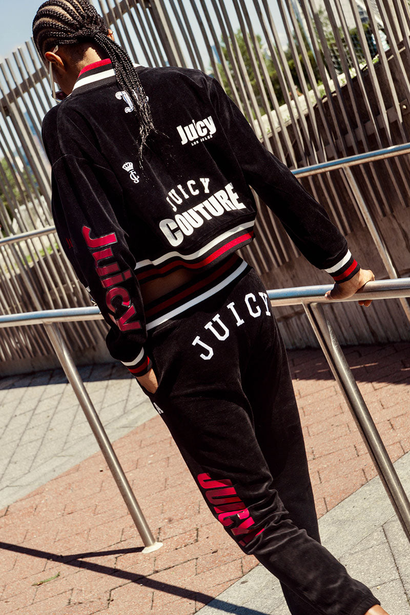 A person with braided hair wears the Mixed Logo Cotton Velour Jogger by Juicy Couture—a black tracksuit featuring bold white and pink logos on the back, arm, and leg—as they walk outdoors along a metal railing on a sunny day. The iconic cotton velour joggers make this sporty staple effortlessly stand out.