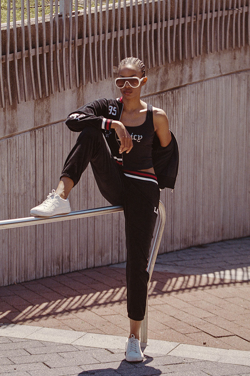 A person wearing sunglasses, a black tracksuit featuring the Mixed Logo Cotton Velour Jogger and white sneakers leans against a metal railing outdoors, with one leg raised and their arm resting on their knee. A concrete wall and fence are in the background.
