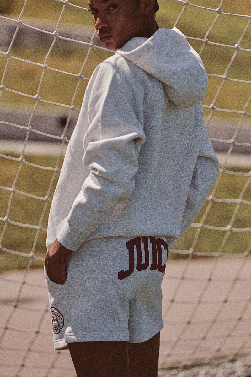 A person with short hair, wearing a heather gray Juicy Couture hoodie and matching Scottie Fleece Booty Shorts, stands outdoors in front of a soccer net, looking over their shoulder at the camera. The shorts feature a large maroon JUICY logo on the back.