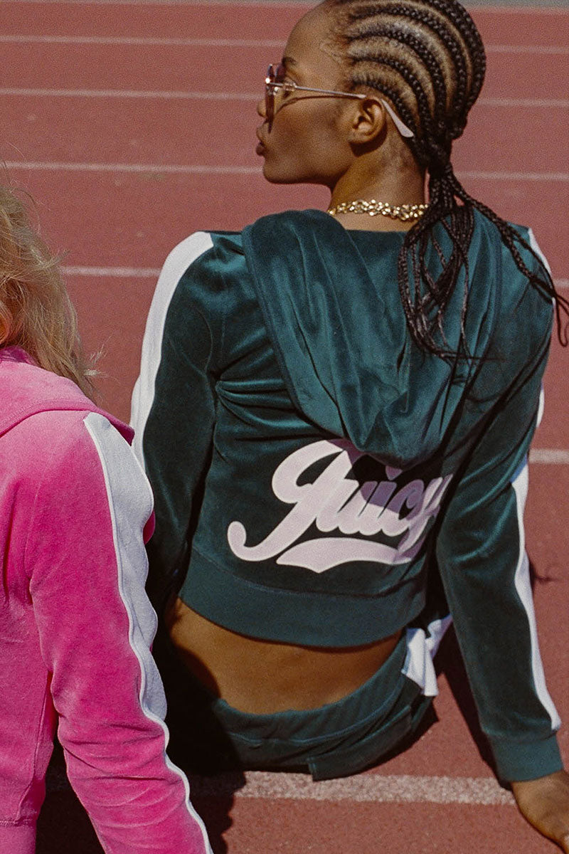 Two people sit on a running track. The person in the foreground wears the Cotton Velour Hoodie, which is dark green with the signature script logo "Juicy" on the back, sunglasses, and has cornrow braids. Another person in a pink tracksuit is partially visible.