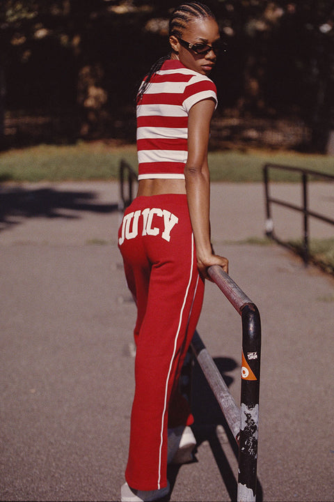 Wearing Scottie Fleece Wide Leg Pants in red with the Juicy logo and a red-and-white striped crop top, this person stands outdoors, holding onto a railing and looking back over their shoulder while wearing sunglasses.