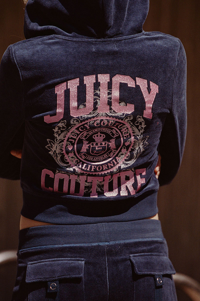 Wearing the Scottie Cotton Velour Hoodie by Juicy Couture in dark blue cotton velour, featuring a large pink "Juicy" and a decorative Scottie crest logo printed on the back.