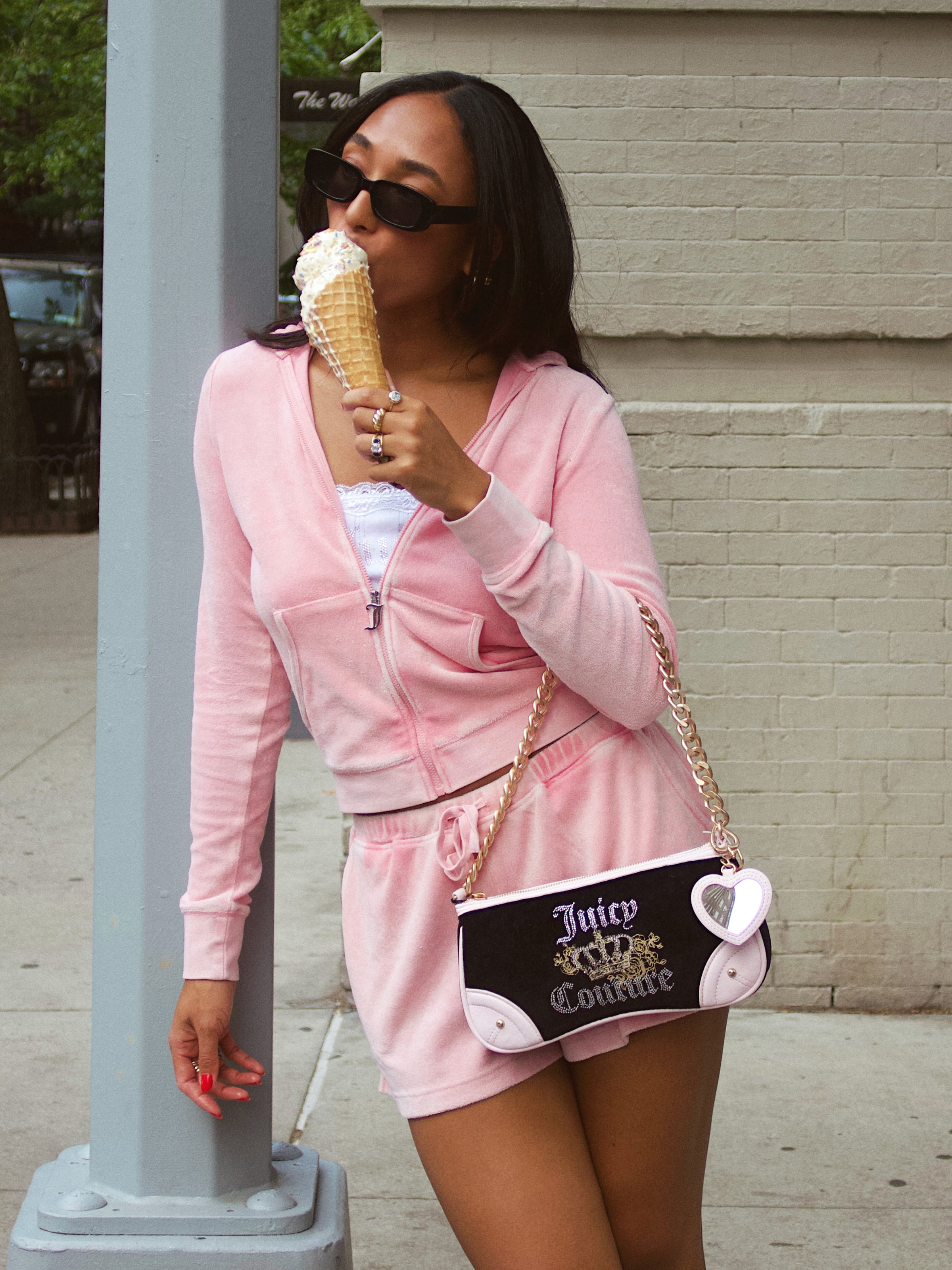 A woman in a pink hoodie and shorts holds ice cream, her Royal Bloom Shoulder Bag bringing timeless glamour. Sunglasses on, she leans against a light gray pole on the city sidewalk, displaying effortless style and a sleek silhouette.