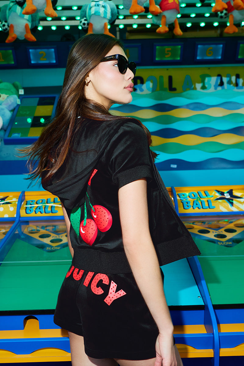 A woman wearing sunglasses and a black outfit with cherries and the word JUICY on the back stands in front of a colorful Roll A Ball carnival game. Bright lights and stuffed animal prizes are visible in the background.