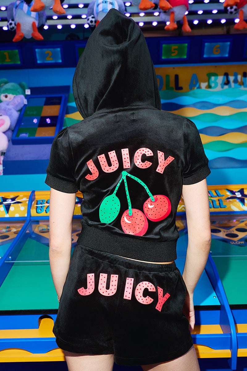 A person wearing a black velour hoodie and shorts with “JUICY” and cherry graphics on the back stands in front of a colorful arcade skee-ball game. Their hood is up, and stuffed animals are visible in the background.