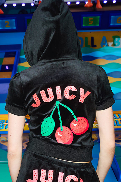 Wearing the Cherry Bling Velour Hoodie—a black, short-sleeve hoodie with rhinestones and a Juicy cherry graphic on the back—a person stands in front of a vibrant carnival game display.