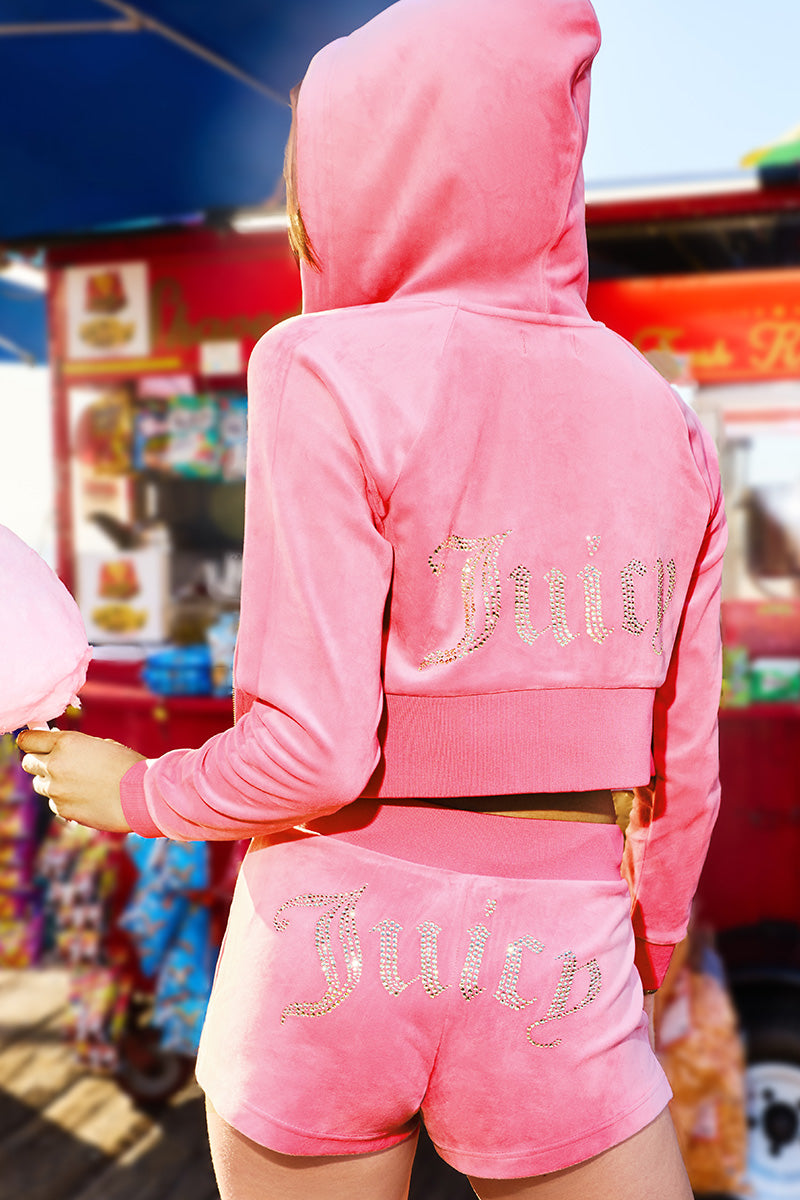 Someone stands outdoors by a colorful food cart, dressed in the Scottie Big Bling Velour Hoodie and matching shorts, featuring a Juicy bling logo.
