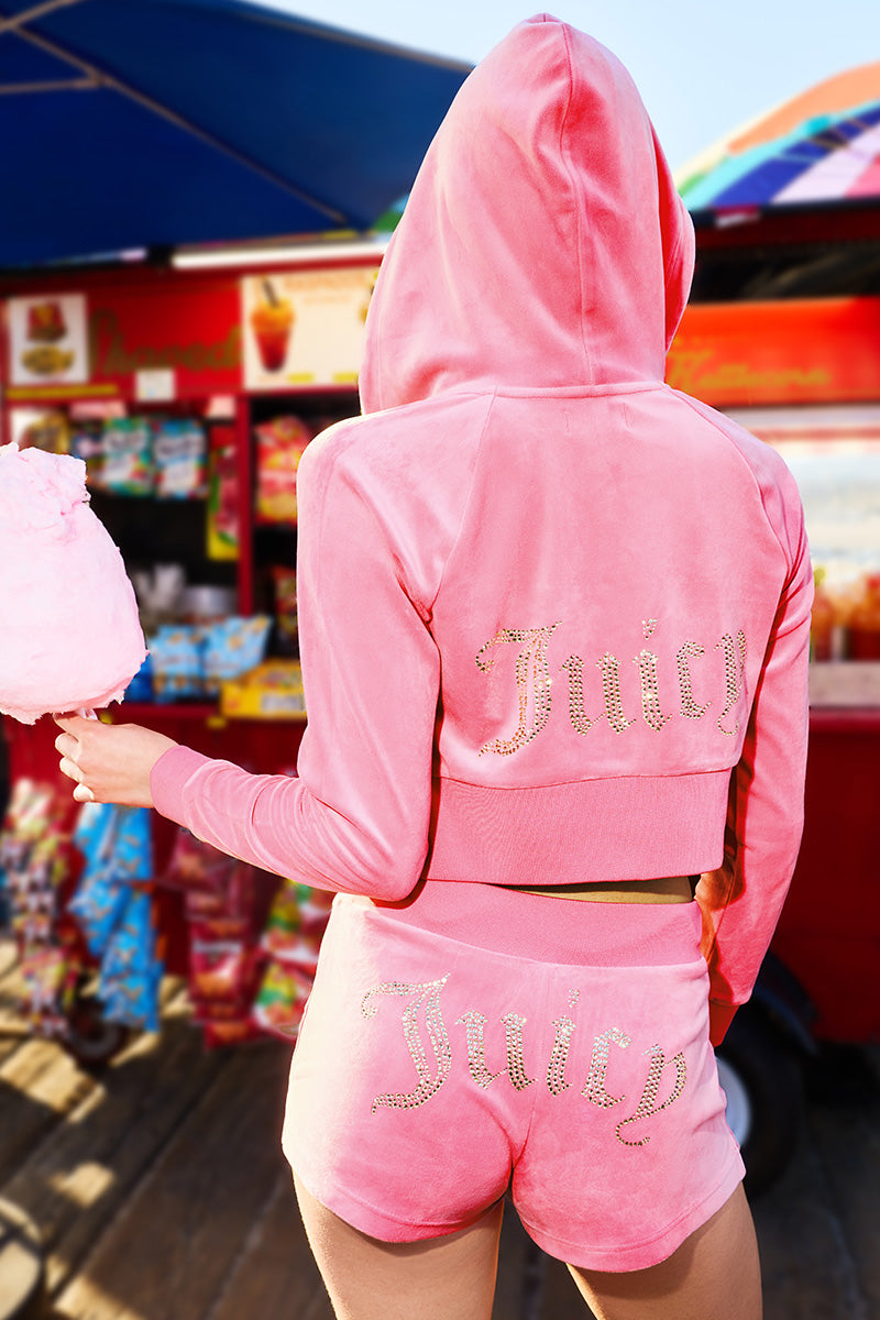 A person faces away, wearing the Scottie Big Bling Velour Hoodie in pink with a Juicy logo and zip front, holding pink cotton candy in front of a colorful snack stand.