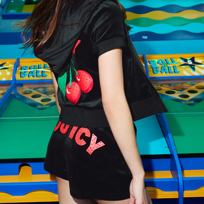 A person in a black velour hoodie and shorts with red JUICY lettering and a cherry graphic stands in front of a colorful arcade skee-ball game.