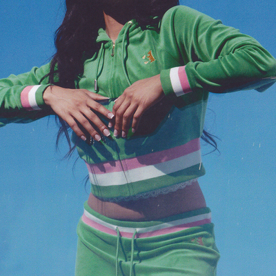 A person wearing a green velour tracksuit with white and pink stripes poses against a clear blue sky, with their hands held up near their chest. Their face is partially out of the frame.