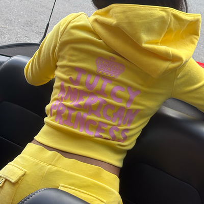 A person sitting in a car, wearing a bright yellow hoodie and pants set. The back of the hoodie reads JUICY AMERICAN PRINCESS in large pink letters under a pink crown graphic.