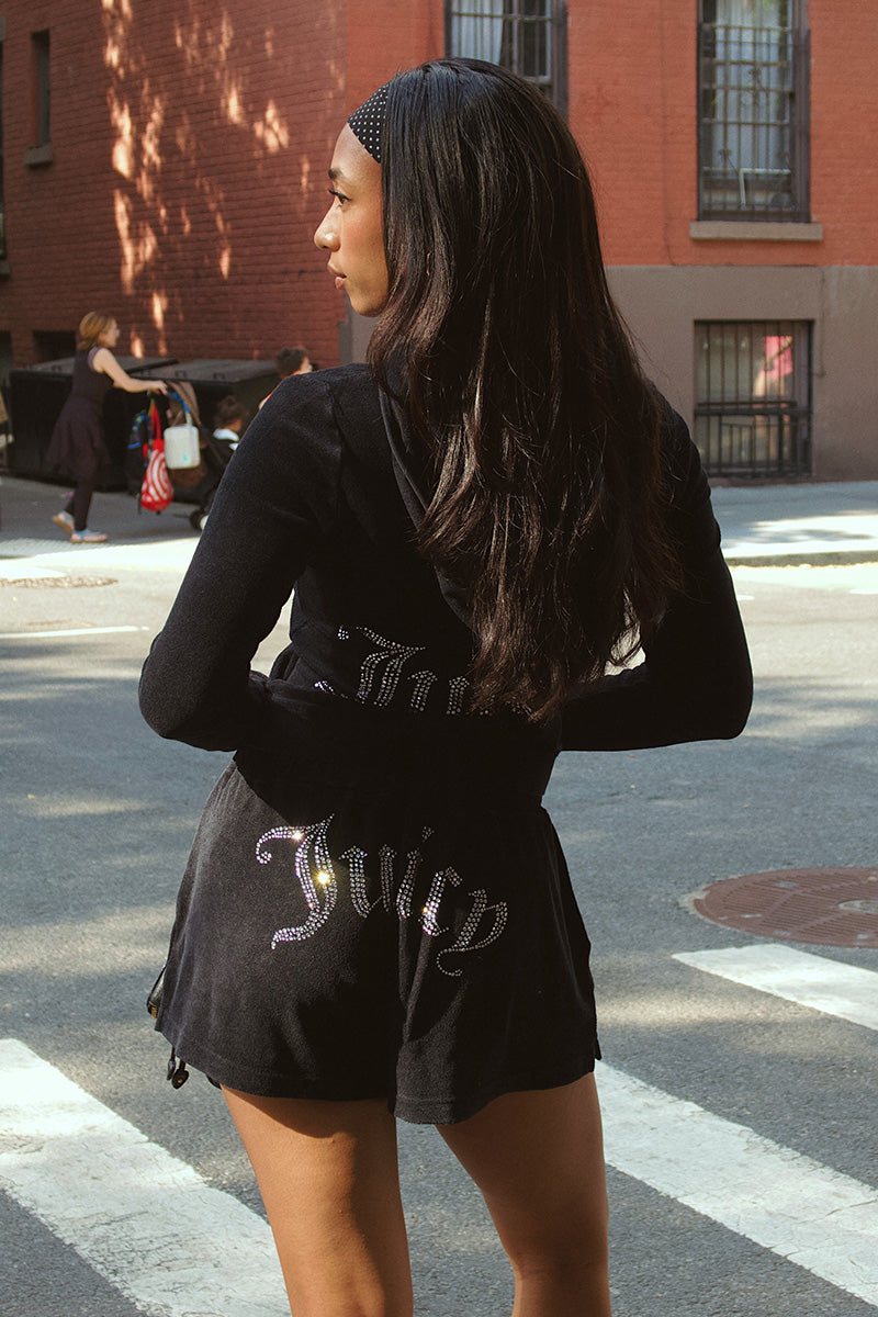 A woman with long dark hair stands at a crosswalk, facing away. She's wearing a black headband, black top, and Big Bling Washed Terry Shorts featuring a rhinestone logo on the back. A red brick building is in the background.