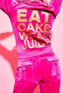 Person wearing a bright pink tracksuit with gold text reading "Eat Cake Wear Juicy" on a pink background